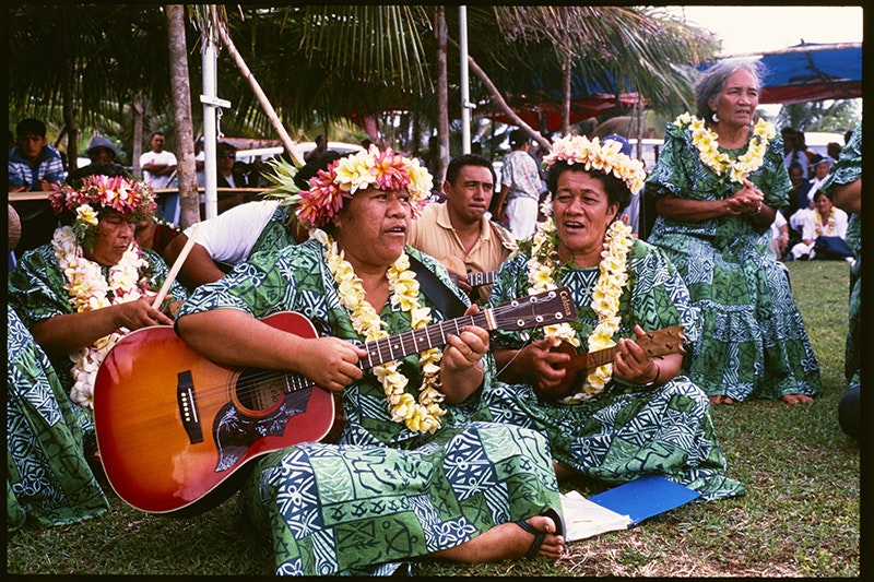 Women wearing the same dresses and headwear are sitting in a group. One is playing the guitar and one is singing.