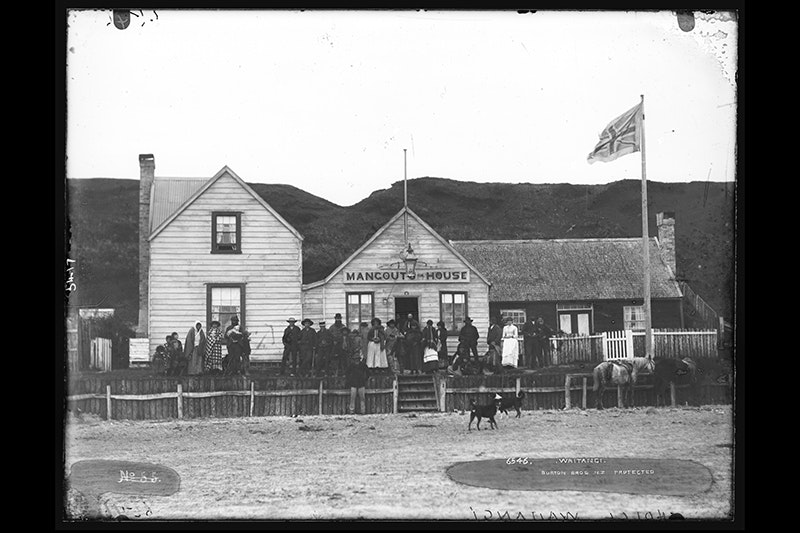 A very old photo with people standing in front of a wooden building. There is a dog in the foreground.