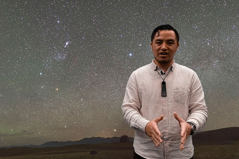 Te Papa staff member Hauiti Gardiner talks to the camera. Behind him is a photograph of the night sky