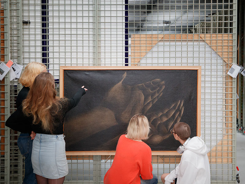 Group of teenagers look at a painting hanging on a rack in the painting store room