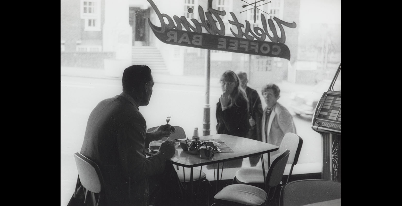 Man sits at a table alone in a cafe, looking out the window, while three people outside look in