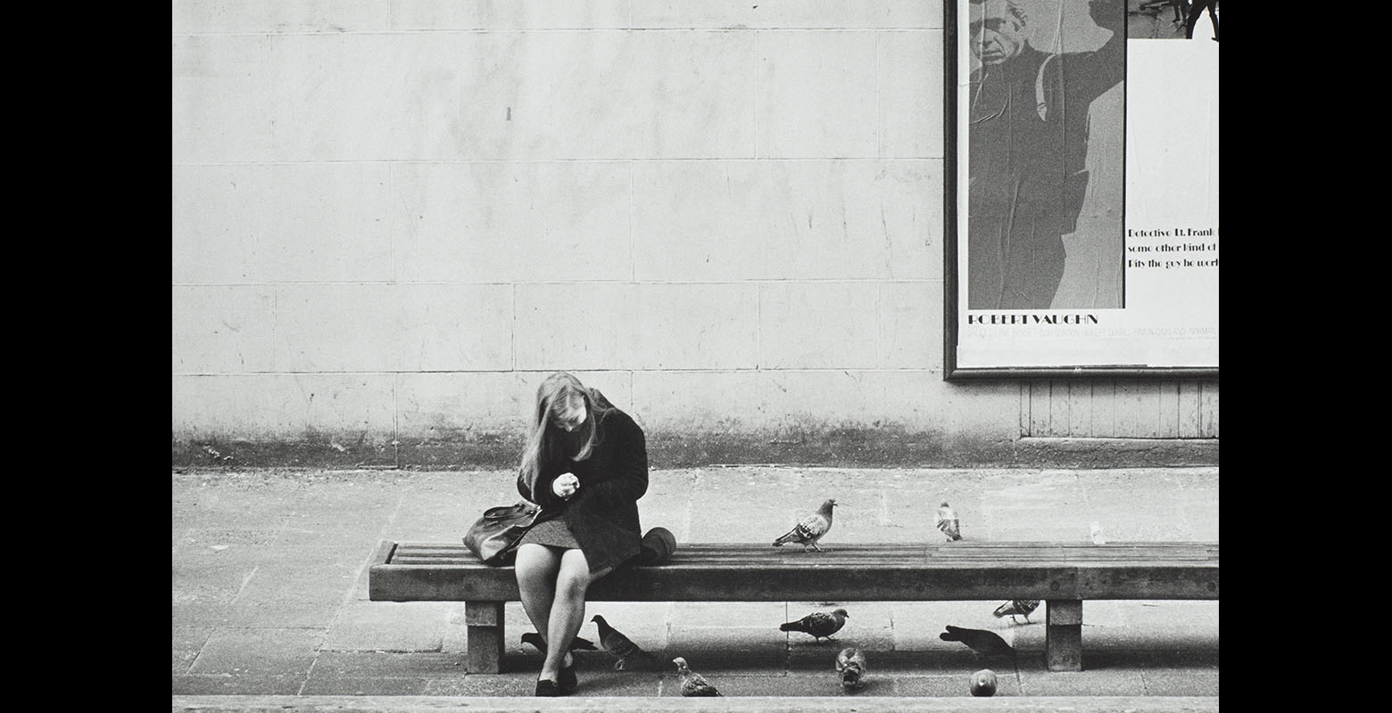 Woman sits on a bench surrounded by pigeons. Behind her on a wall is a poster for a film