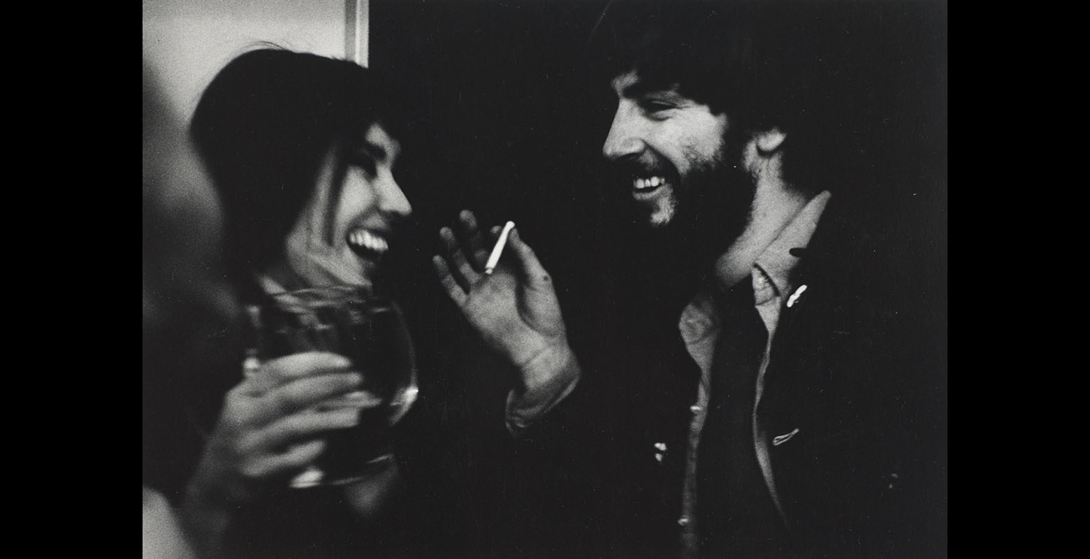 Two people laugh and smile in conversation. One is holding a glass and the other is holding a cigarette