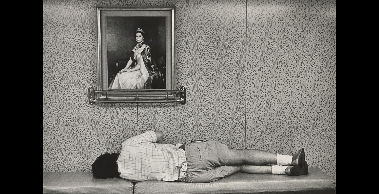 A man lies on a bench, his back to the camera. There is a portrait of Queen Elizabeth the second on the wall above him