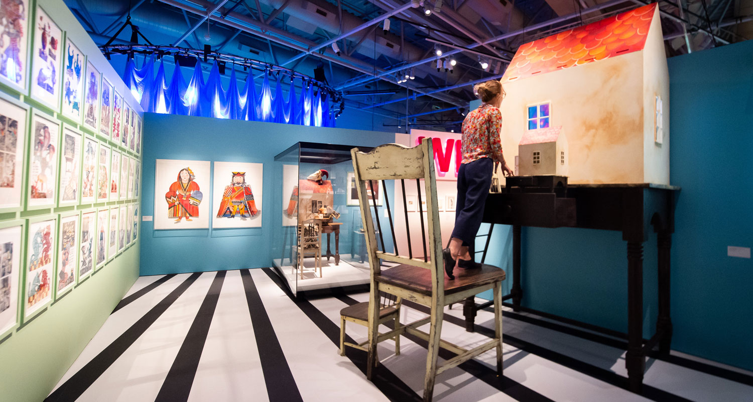 A person stand on a giant chair and peers into a dolls house