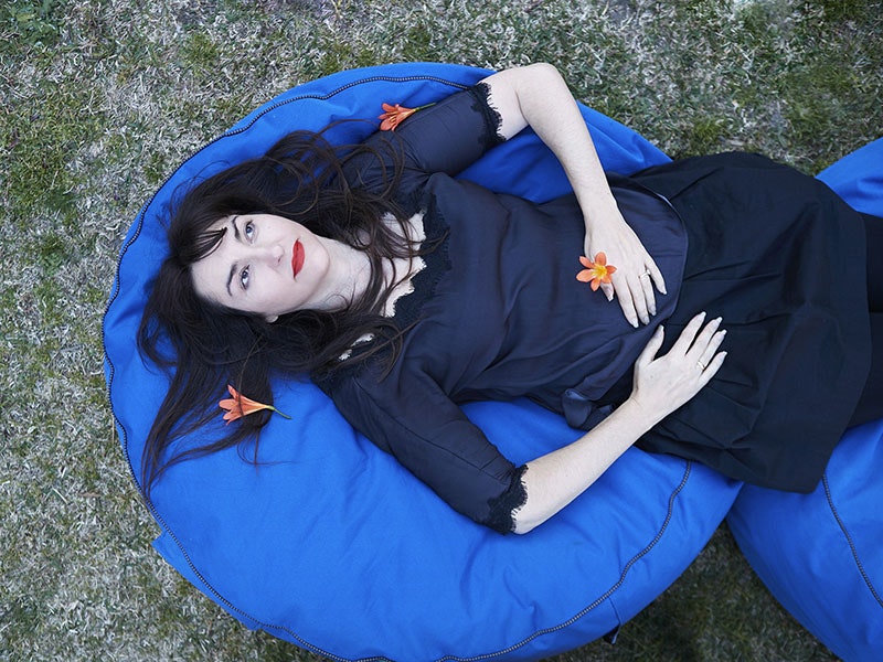 Nike Savvas, dressed in a dark blue dress, lies on a lighter blue beanbag on the grass, holding and surrounded by orange flowers