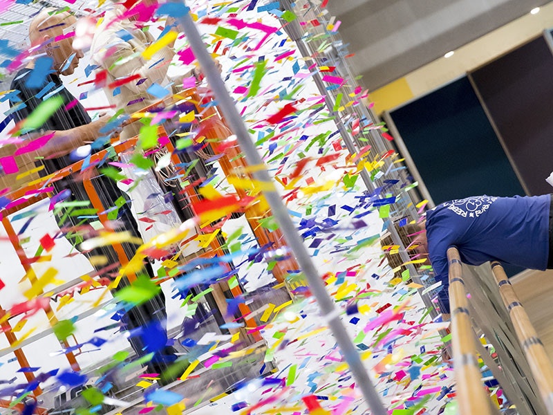 A person on a scissor lift is obscured by a dense collection of strands of colourful pieces of plastic hanging from the ceiling while another person looks over a bridge beside the work down towards the floor, where the work continues
