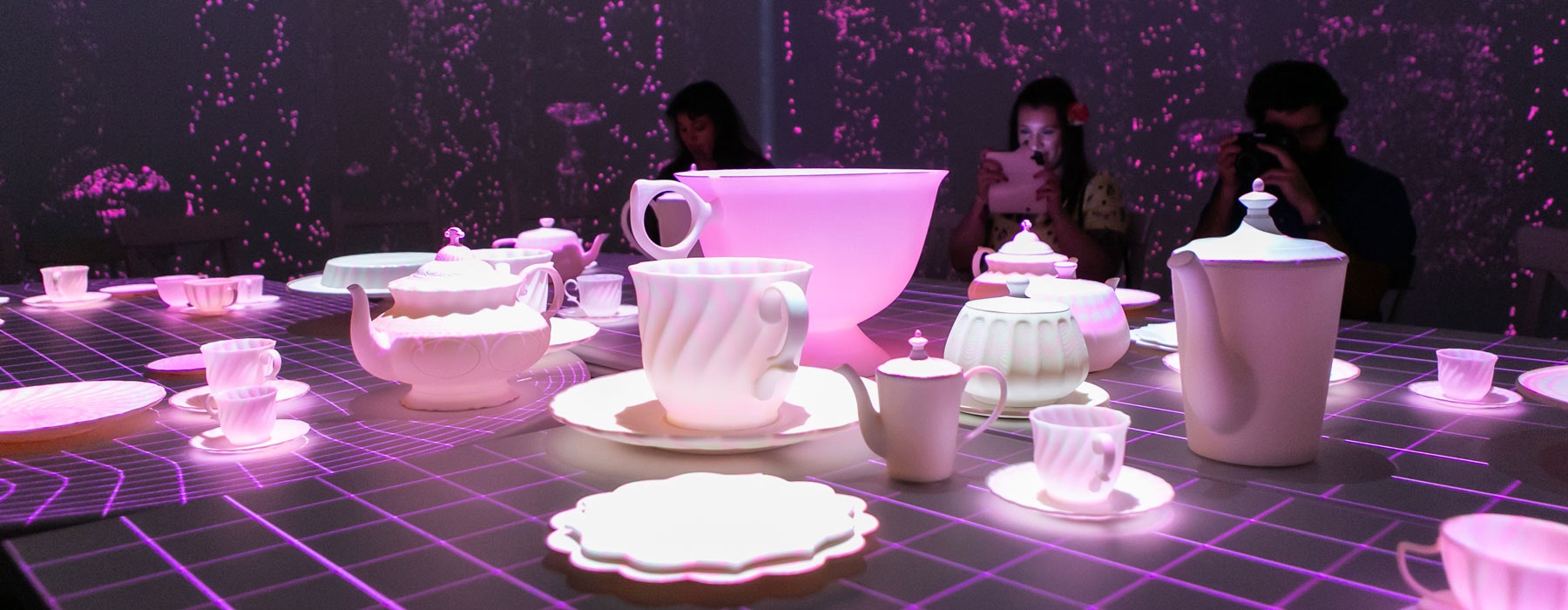 Two people sit at a table with a tea set on it. In the background are projected images of mushrooms and trees