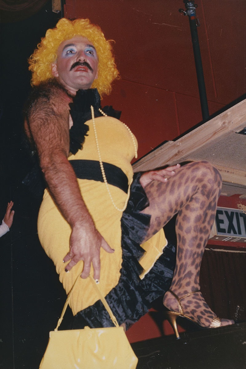 A man in a yellow dress with black highlights and wearing a yellow wig shows off his leg featuring leopard skin tights.