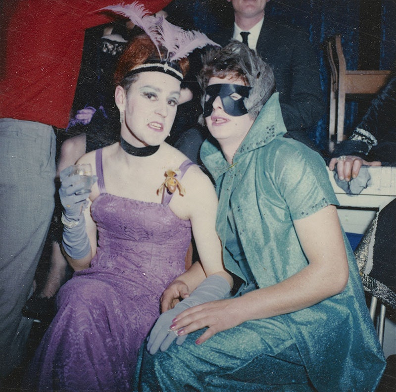 Two people pose for a photo: one in a purple dress and overall Weimar-era look, the other in a green two-piece dress and cape with black mask with feathers covering their eyes