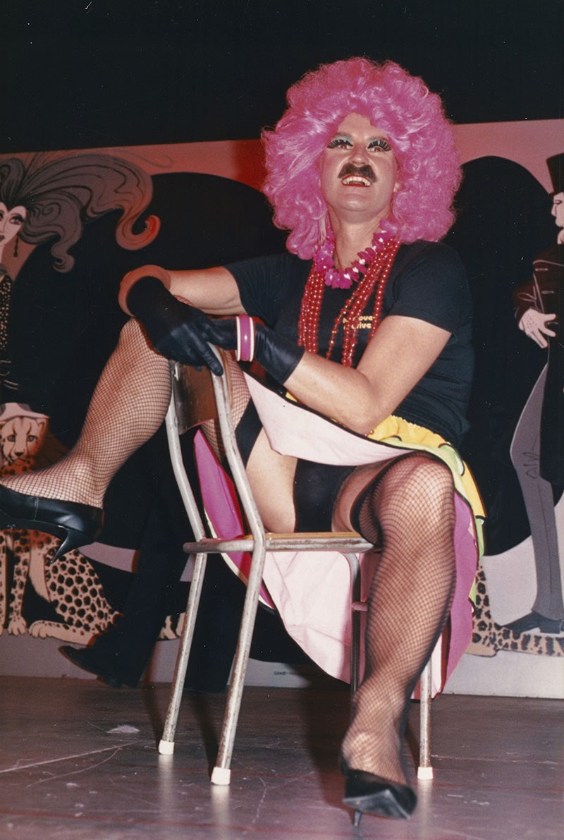 A man in a pink wig, black T-shirt, and dress poses on a chair with his legs open, revealing his underpants.