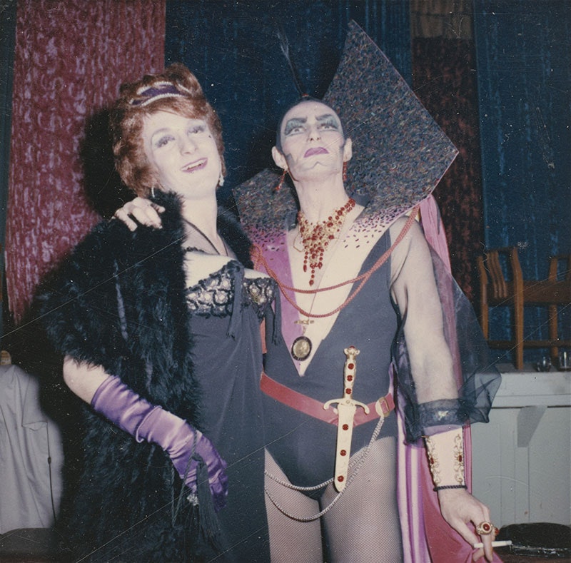 Two people are wearing elaborate costumes: one in old fashioned ball gown attire and the other in a futuristic leotard and cape ensemble