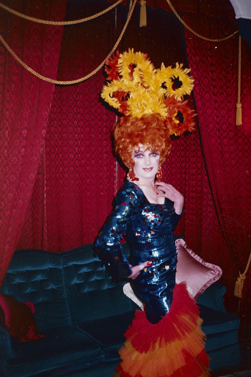 Man dressed in dark blue dress with sequins and puffy red and orange bottom, with an orange wig and bright sunflowers in his hair