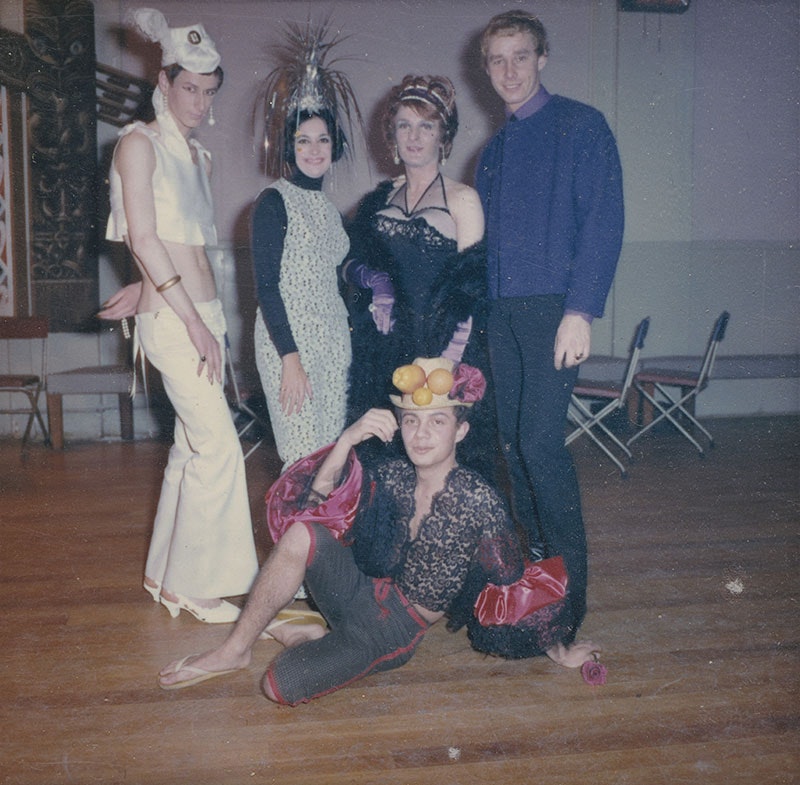 Five people pose for a photo. Four stand while one person sits on the ground. They are in a hall