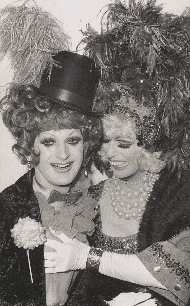 Two men dressed in drag. One wears a top hat and suit with large frilly cravat with a flower on their chest. The other is in a dress with pearls and ostrich feathers in their hair. They are touching the other person's bosom.