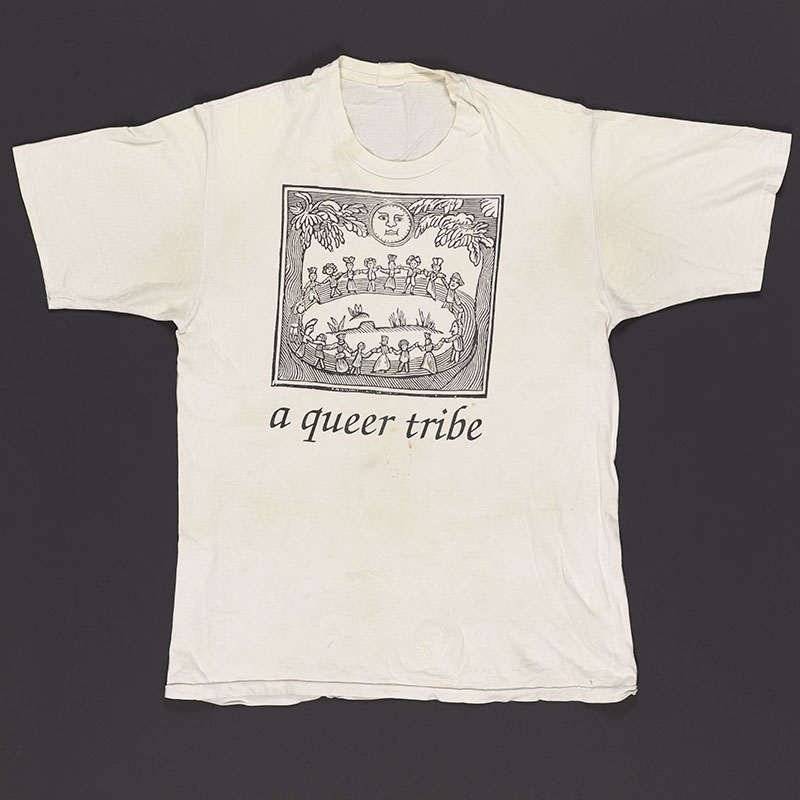 White T-shirt with a graphic in linocut style of people holding hands in a circle while the moon shines above. The words 'A queer tribe' are written below