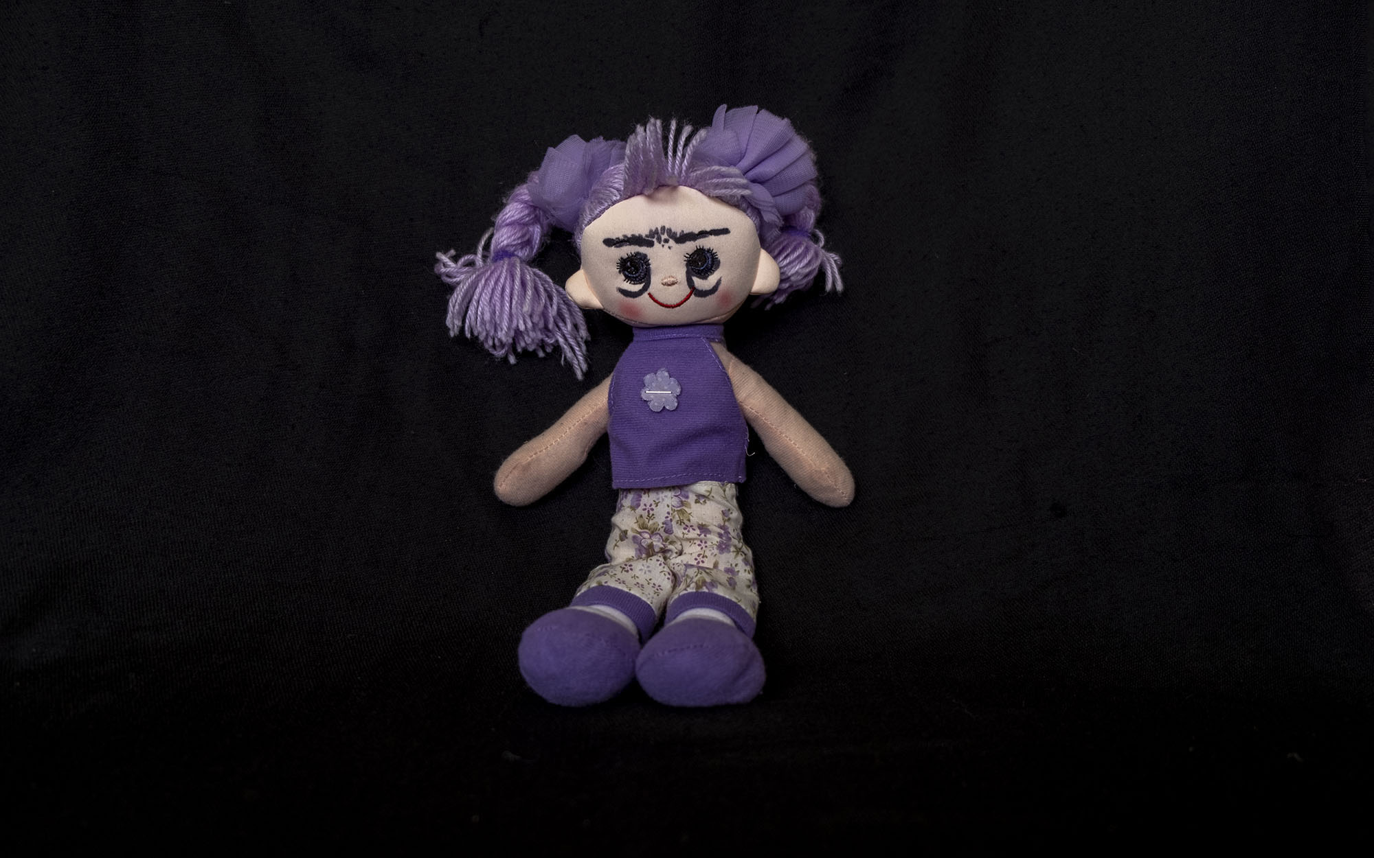 Smiling plush doll with purple hair, a purple singlet, purple shoes, and cream pants with purple flowers and hem