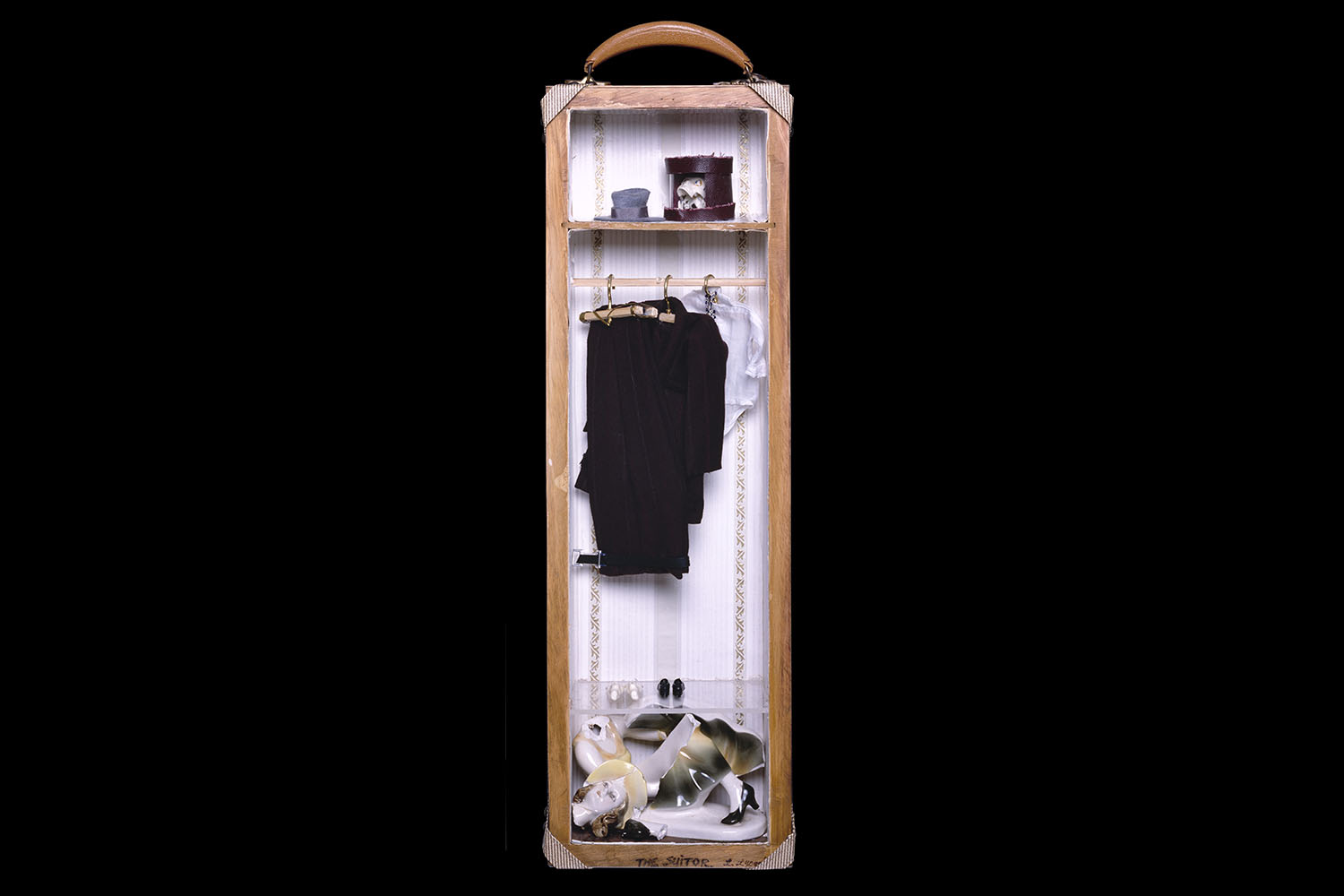 Narrow open half of a suitcase that resembles a wardrobe, inside containing a broken statue of a woman, clothes hanging, and a skull in a hat box