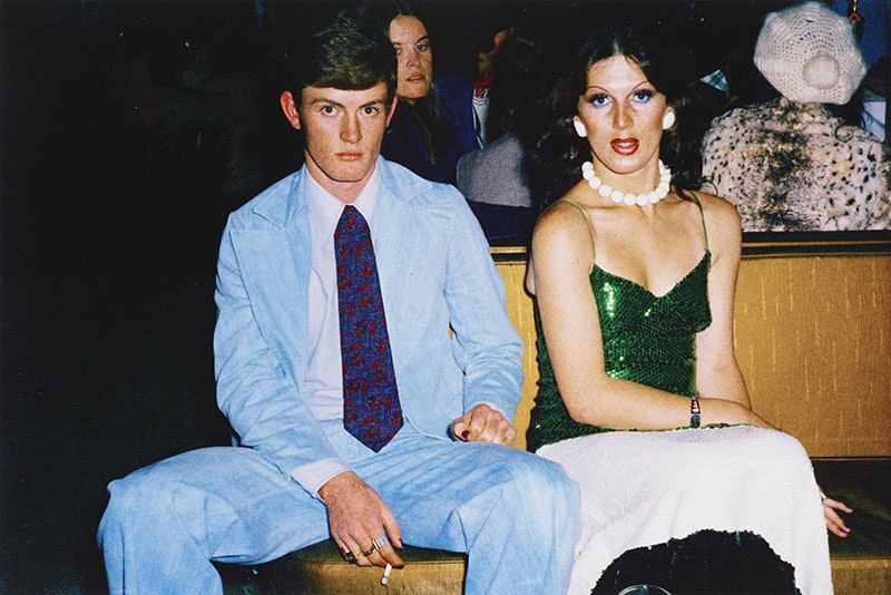 Man in light blue suit and a blue and red tie smoking a cigarette sits beside a man in drag wearing a green sequin dress and a necklace made up of large white balls