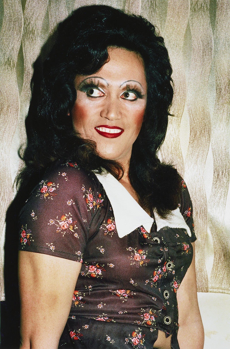 Man in drag wears a dark-coloured dress, with white collar, decorated with small floral bouquets