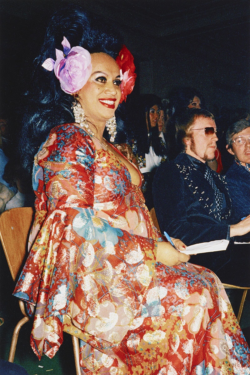 Carmen sits in a chair with a notebook and pen, wearing a predominantly red floral dress and flowers in her hair