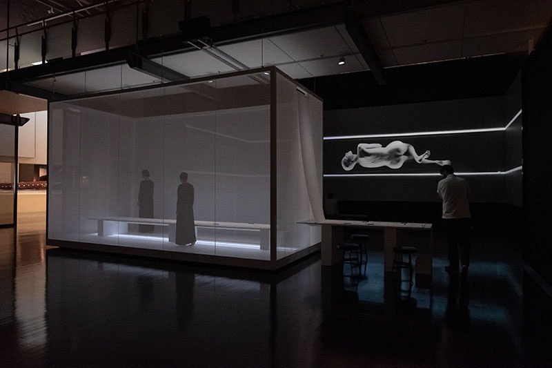 A man walks around a space that contains a box with two women inside and a massive projection on the wall of a woman contorting her body