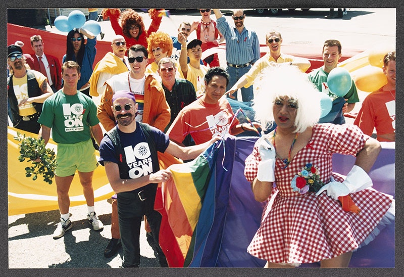 Group of people in an assortment of of costumes stand in a parade holding the rainbow flag