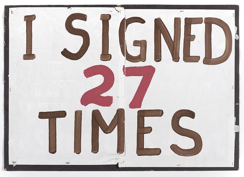 Cardboard sign painted white with the words ”I signed 27 times” on it