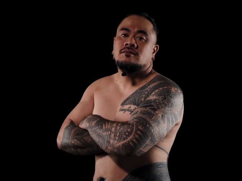 A man with Sāmoan tattoos stands with his arms crossed