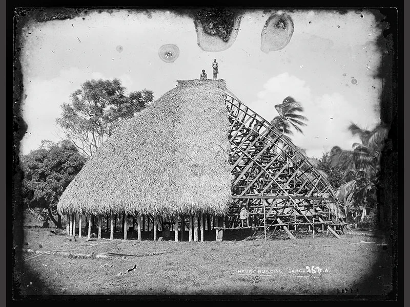 Several unidentified men are standing on the framework of a partially-thatched faletele, grass in foreground and trees and palms in the background
