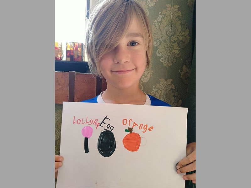 Boy holds sheet of paper. On the paper he has drawn a lollipop, and egg, and an orange to represent the letters of his name