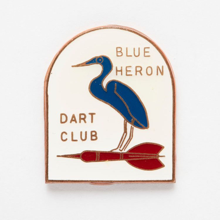 Badge in the shape of an archway featuring an illustration of a blue heron and a red dart with the words “Blue Heron Dart Club”