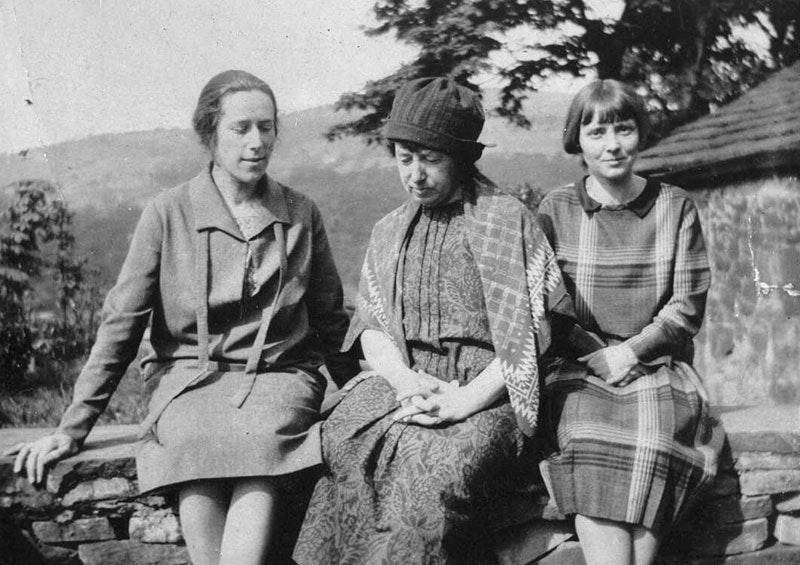 Three women sit on a low brick fence, with a farmhouse-style building behind them to the right. Frances Hodgkins is in the middle, looking somewhat despondent