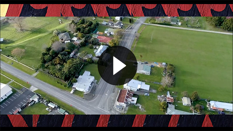 Still from video showing overheard shot of a town, the video footage is frames by carving of a Māori motif