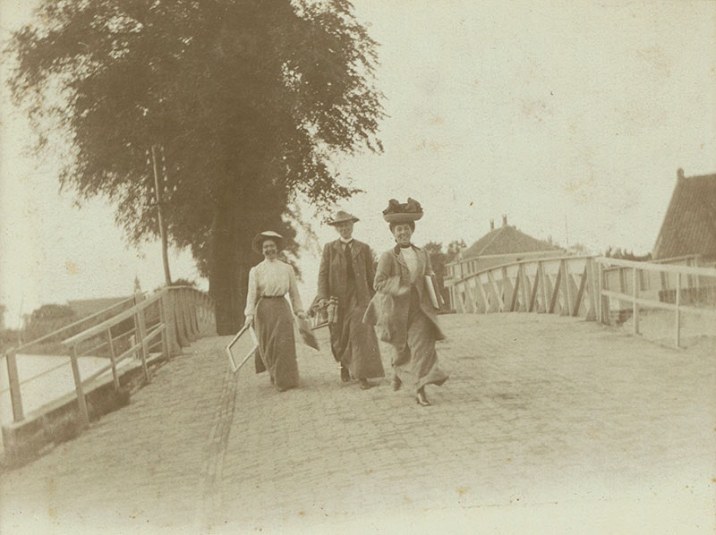 Three woman crossing a bridge. One of the women is running towards the camera, smiling. Behind them is a large tree and buildings