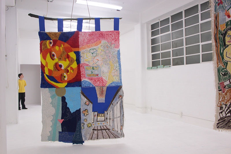 A large quilt hangs from the ceiling of the play_station gallery. It is an almost empty, white space. Part of another quilt can be seen hanging to the right of the frame