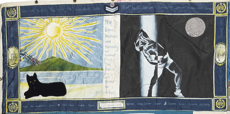 A panel from the NZ Aids Memorial Quilt. A beach scene with a black dog and a mountain in the distance, and a man posing in front of a moon. In the middle are the words ‘The love, humour, and humanity we cannot forget. The friend we will never be without’