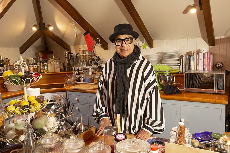Uncle Pare stands in his kitchen. He wear a black and white striped shirt, black scarf, glasses, and a black brimmed hat. His kitchen is overflowing with a well organised array of bottles, utensils, plates, and jars