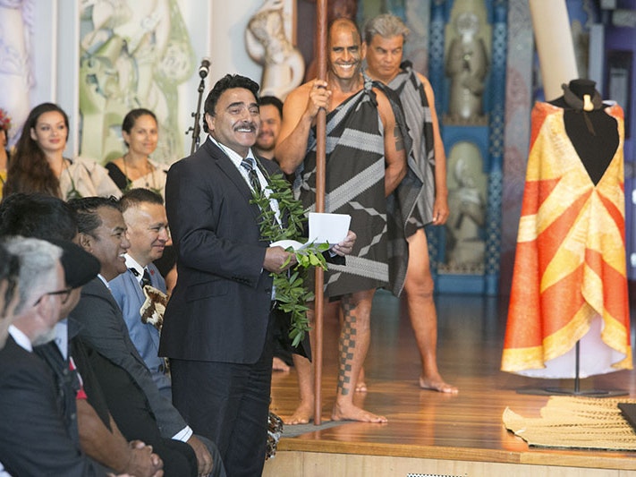 Te Papa Kaihautū Arapata Hakiwai speaks at the ceremony to return the ‘ahu ‘ula and mahiole, 2016. The cloak lies flat while a replica is draped on a mannequin. Te Papa Arapata addresses the room standing in front of a cloak draped on a mannequin