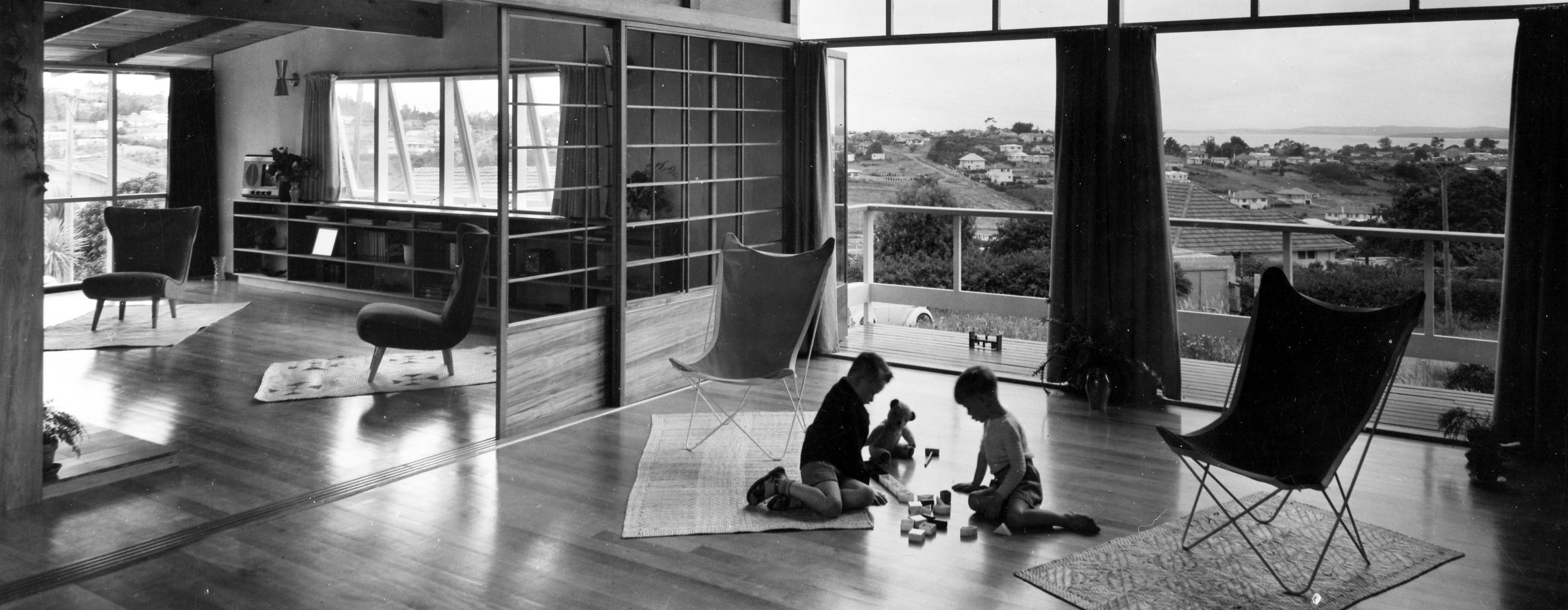Black and white photo of two children playing on the floor of a large room with a view of other houses and the harbour