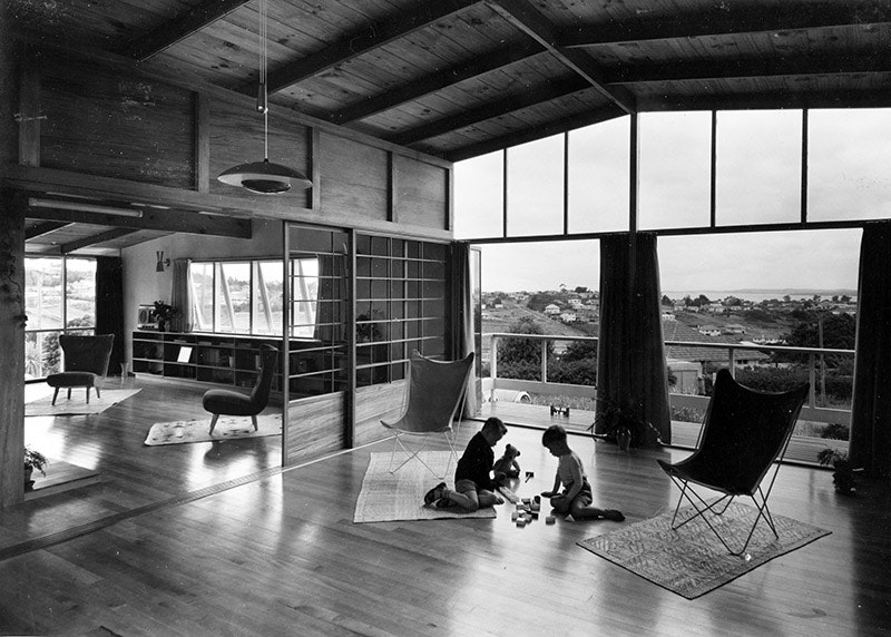 Black and white photo of two children playing on the floor in a big room with a view of other houses and the harbour.