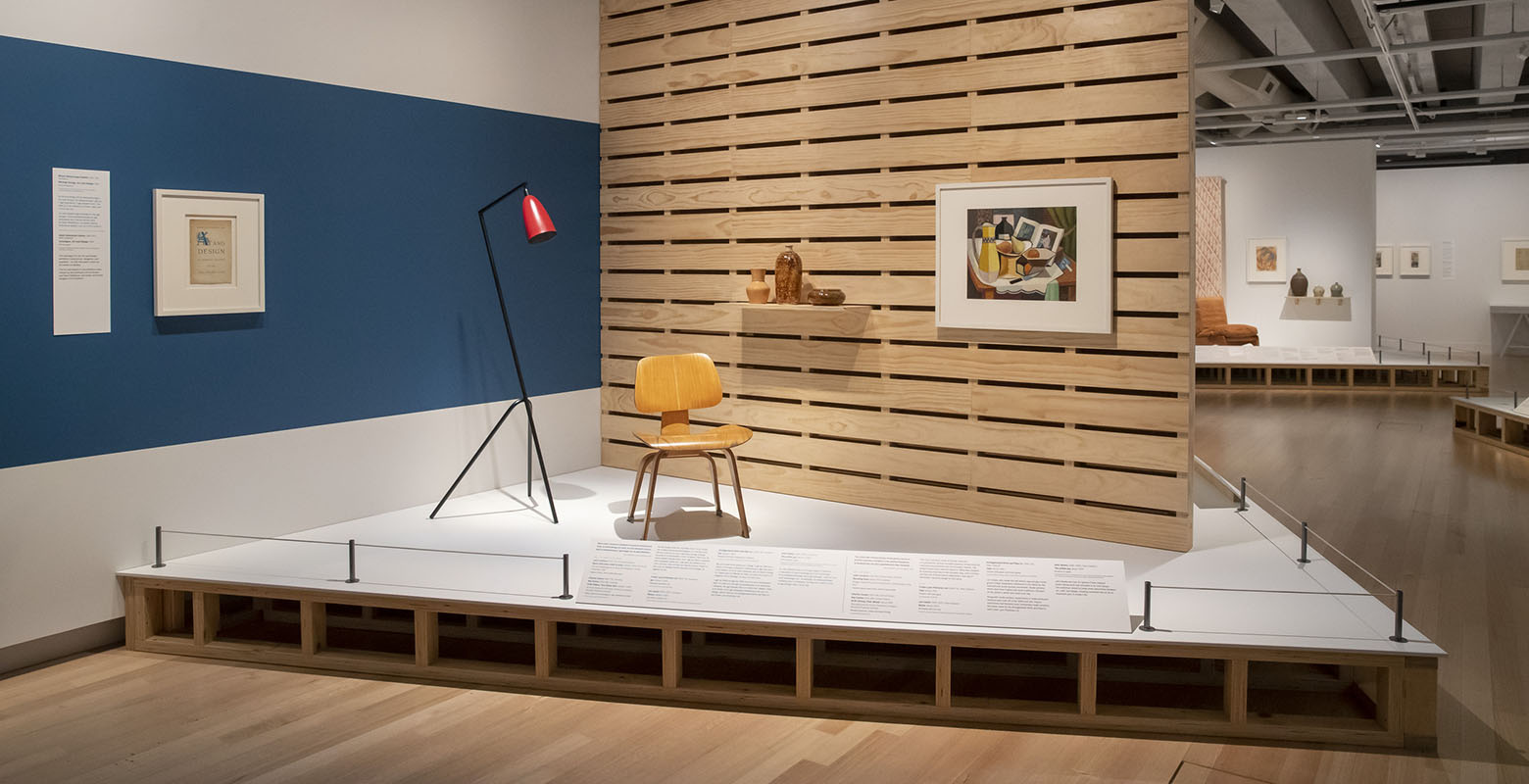 Photo of exhibition view. On a raised platform sits a chair and lamp, with a wooden wall behind. On the wall is a painting of a still life and a shelf with ceramics