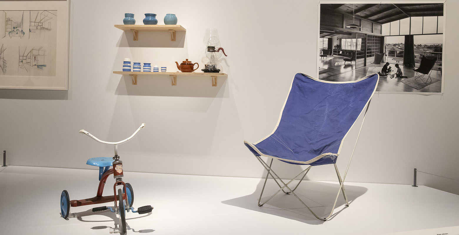Photo of exhibition view. On a raised platform sits a red and blue trike and a blue chair. On the wall is shelving holding a coffee carafe, a tea pot, and a selection of jars. On the wall is a photo of the interior of a modernist house