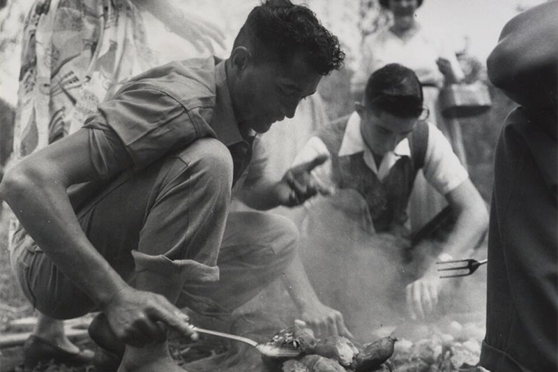 Two young men work to prepare food for a hāngī. The first is using a spatula to check on food from the hāngī while the second is lifting the food from the pit. Three women and another man are in sight around the hāngī pit.