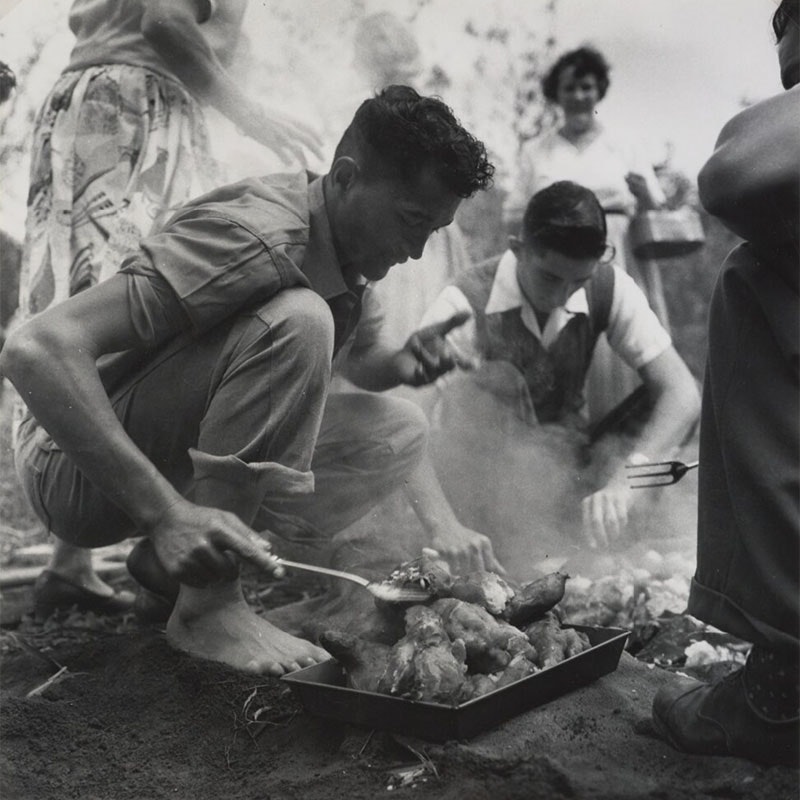 Two young men work to prepare food for a hāngī. The first is using a spatula to check on food from the hāngī while the second is lifting the food from the pit. Three women and another man are in sight around the hāngī pit.