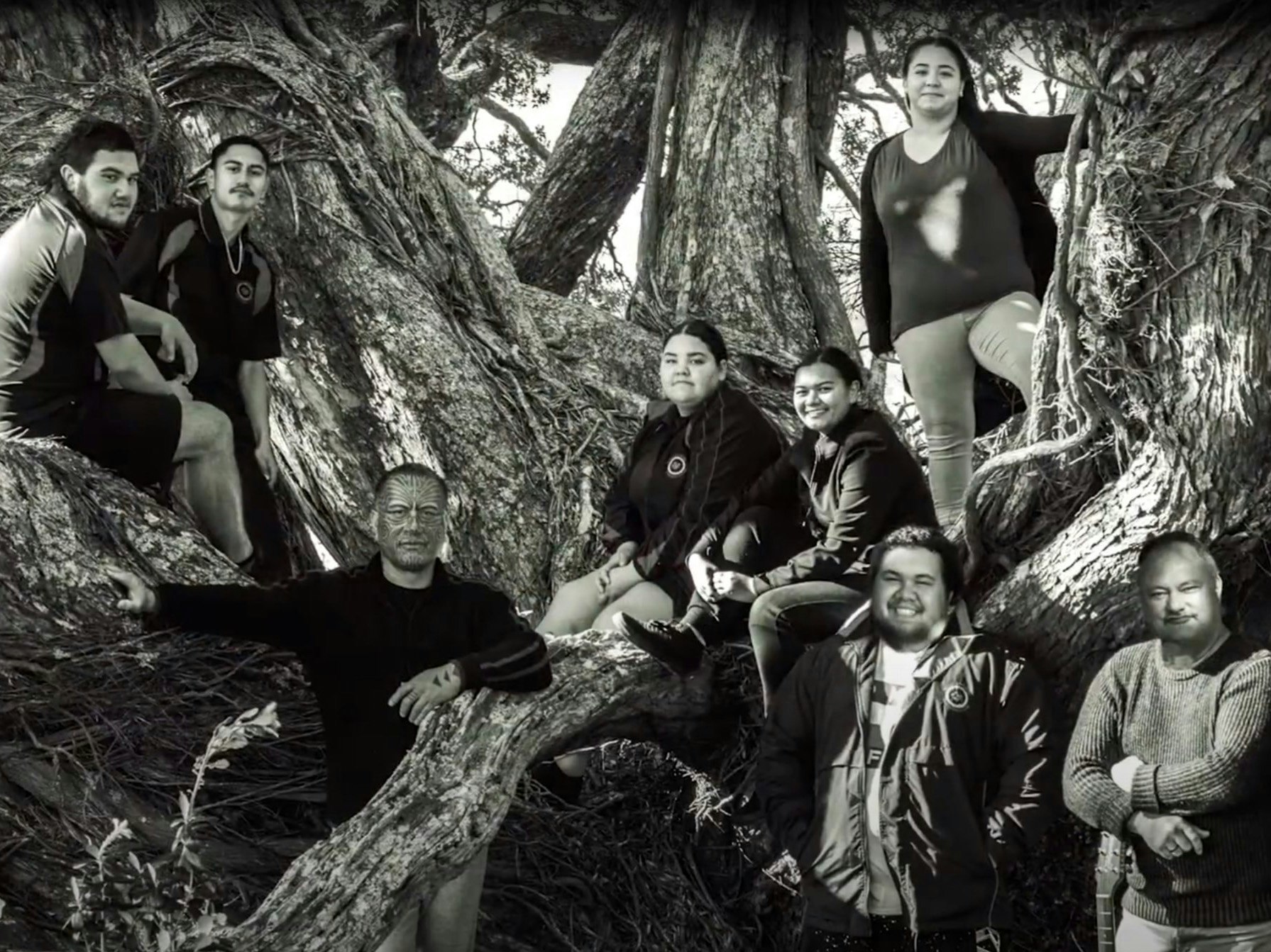A group of people facing the camera sitting in and standing in front of a tree