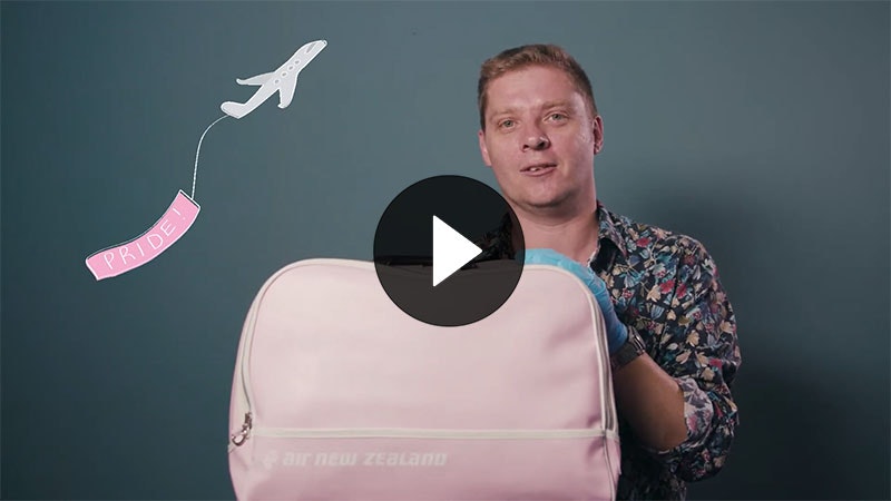 Chris Brickell holds a pink Air New Zealand travel bag. An illustration of an aeroplane flies behind him with a pink flag saying ‘Pride’ behind it