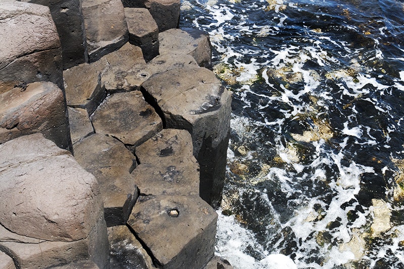 Basalt columns that are mostly five-sided by the sea which has a lot of seaweed and foam in it