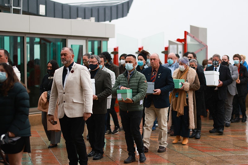 A man in a white jacket leads several people into a building. Many of them are carrying green boxes with human remains in them
