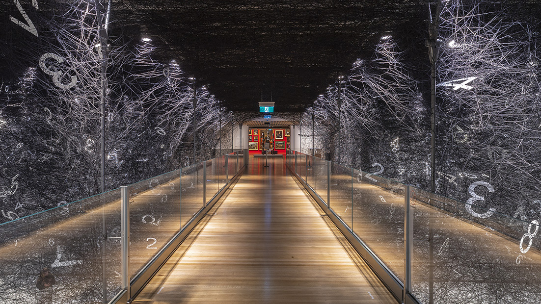 Black wool in a web stretched across a museum gallery floor, with a wooden bridge running through the middle of it. In the distance is a red wall with paintings on it.